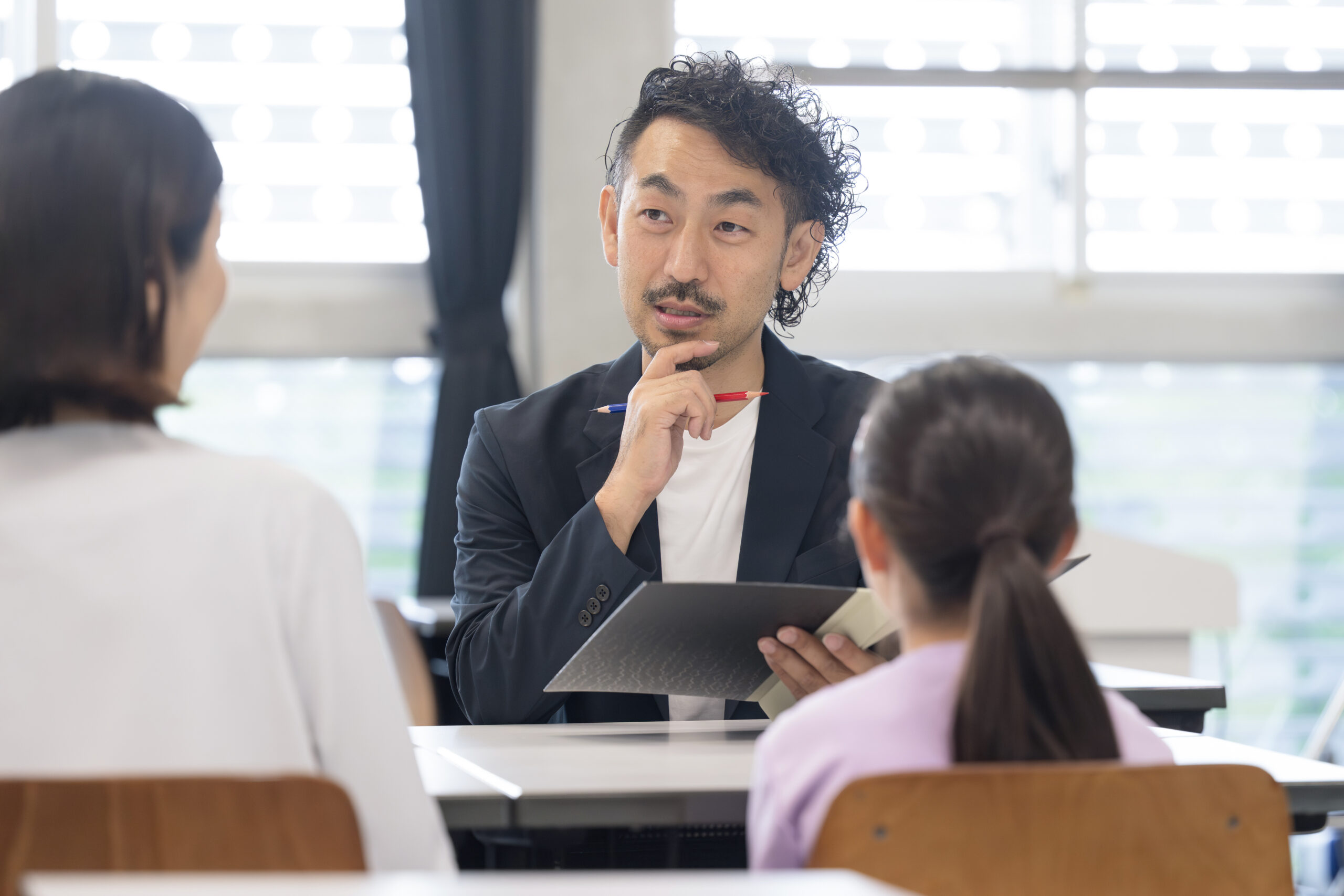 Back of mother and young daughter sitting across a desk during meeting with teacher
